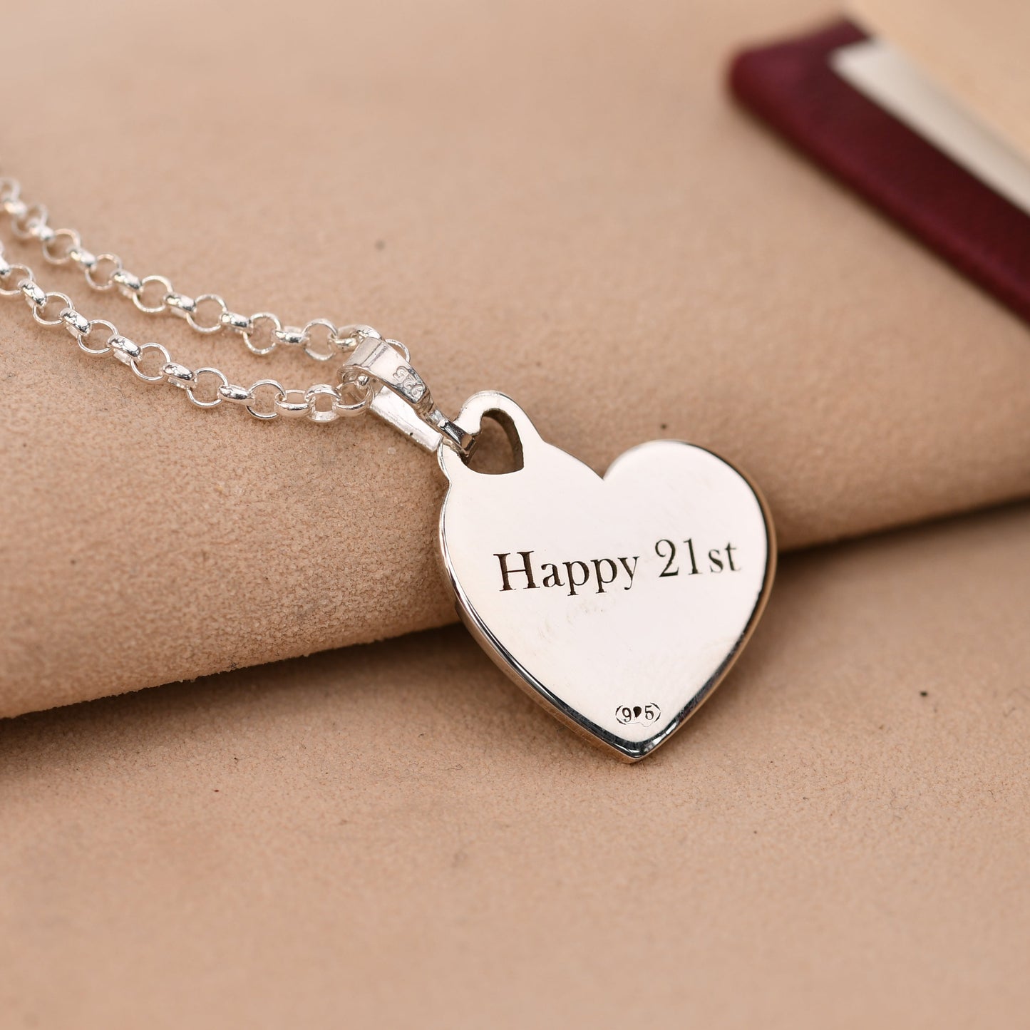 Silver heart-shaped pendant necklace with 'Happy 21st' engraving on a beige surface.