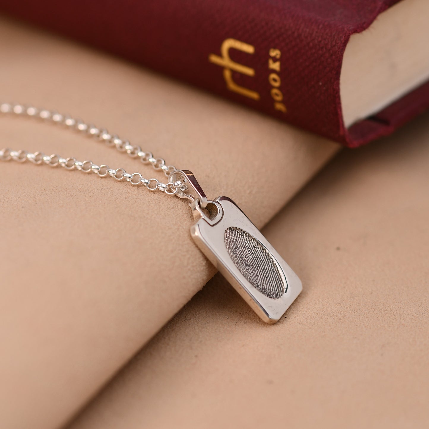 Silver necklace with a fingerprint pendant on a beige surface with a book in the background.