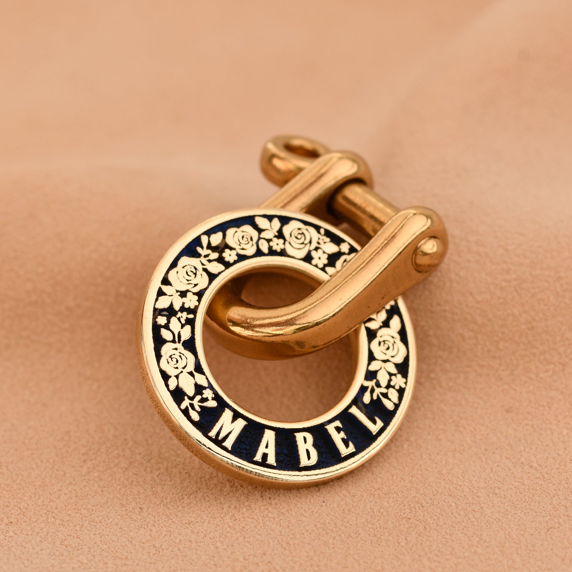 Gold and black enamel pet tag with 'Mabel' branding on a beige fabric background