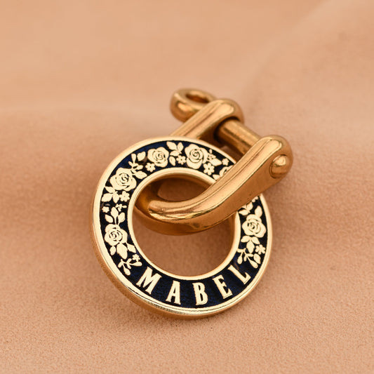 Gold and black enamel pet tag with 'Mabel' branding on a beige fabric background
