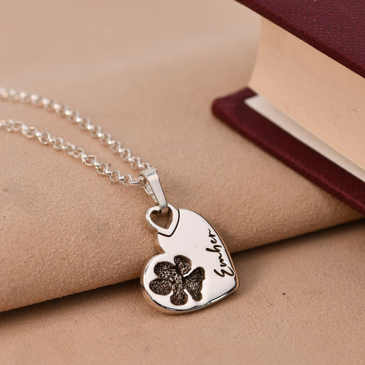 Silver necklace with a heart-shaped pendant featuring a paw print and engraved text on a beige surface.