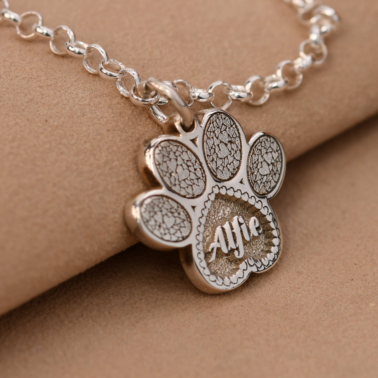 Silver paw-shaped pendant necklace with 'Alfe' engraved on a beige background