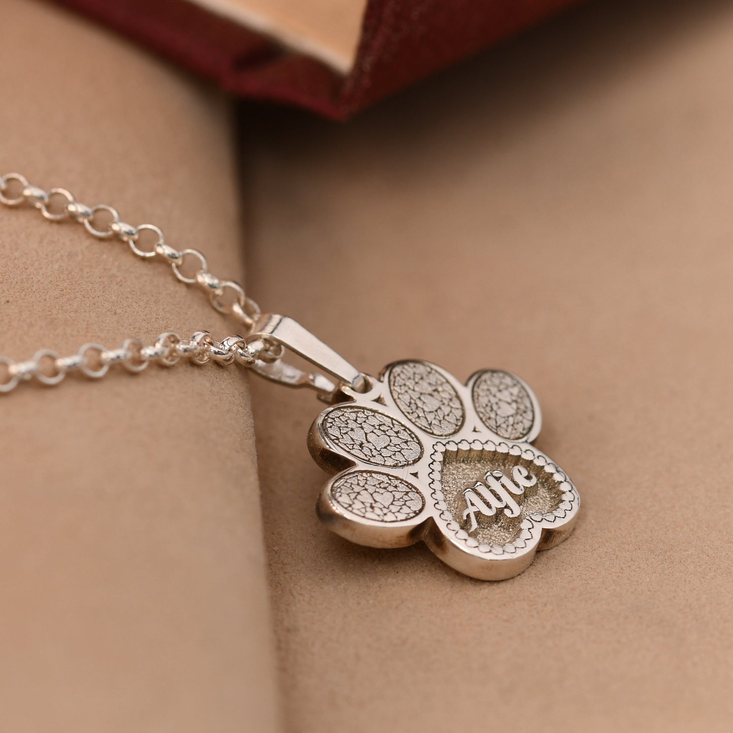 Silver paw-shaped pendant necklace with 'Alfie' engraved on a beige surface
