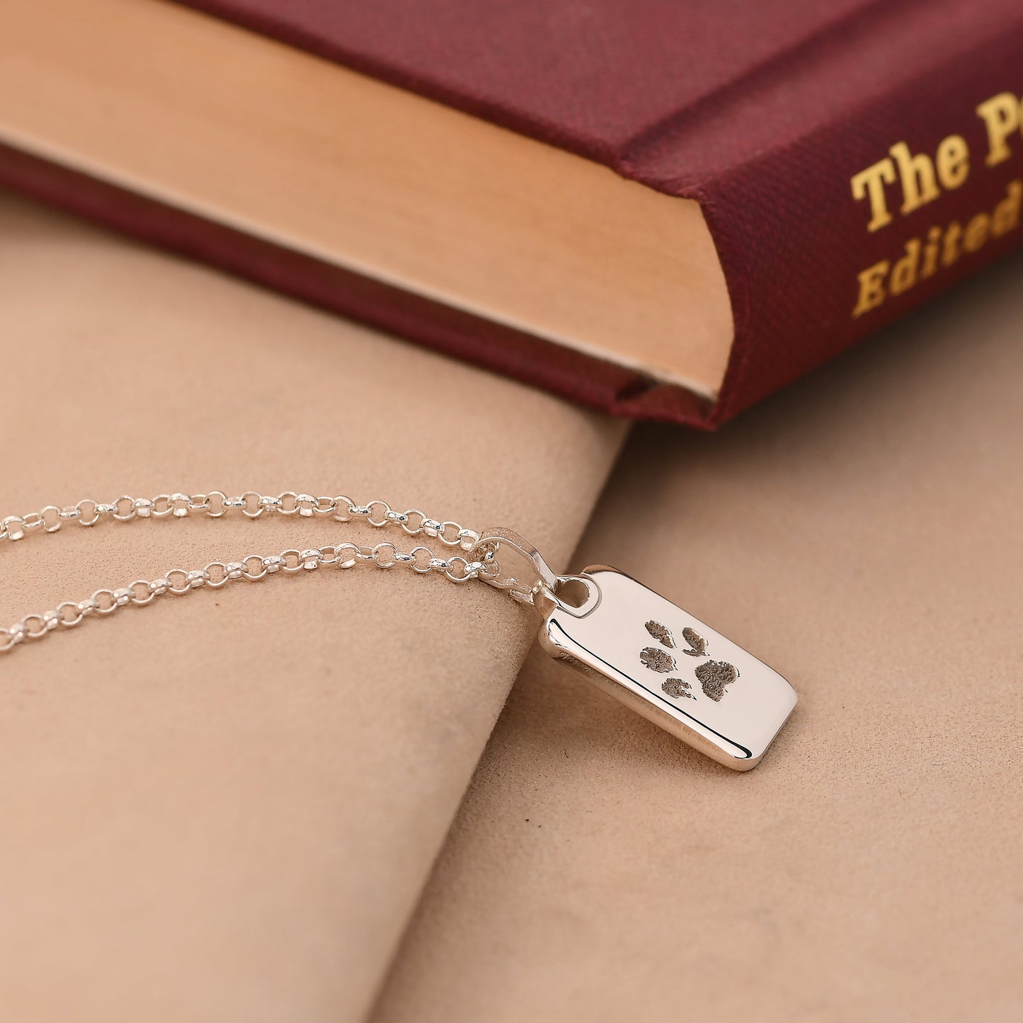 Silver necklace with a paw print tag on a beige surface with a book in the background